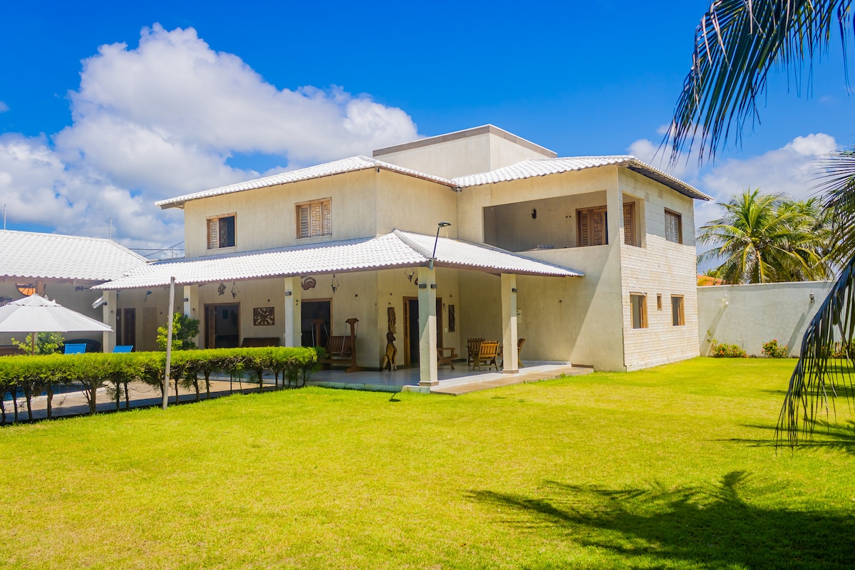 A spacious two-story house is depicted, surrounded by lush green grass. The exterior features a light-colored facade with large windows and a shaded porch. Palm trees line the property, complementing the bright blue sky filled with clouds.