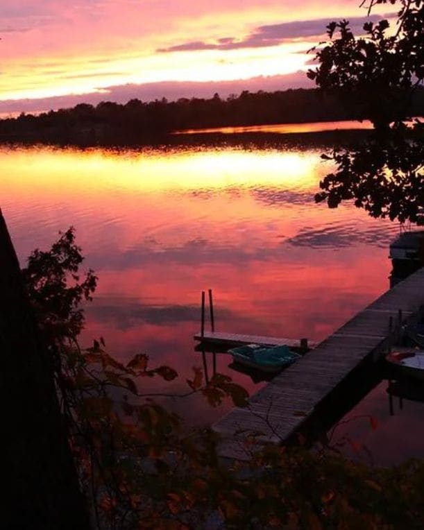 A serene waterfront scene is depicted, featuring a vibrant sunset reflecting in the calm water. Silhouettes of trees frame the image, while a wooden dock extends into the lake, complemented by a small boat positioned nearby.