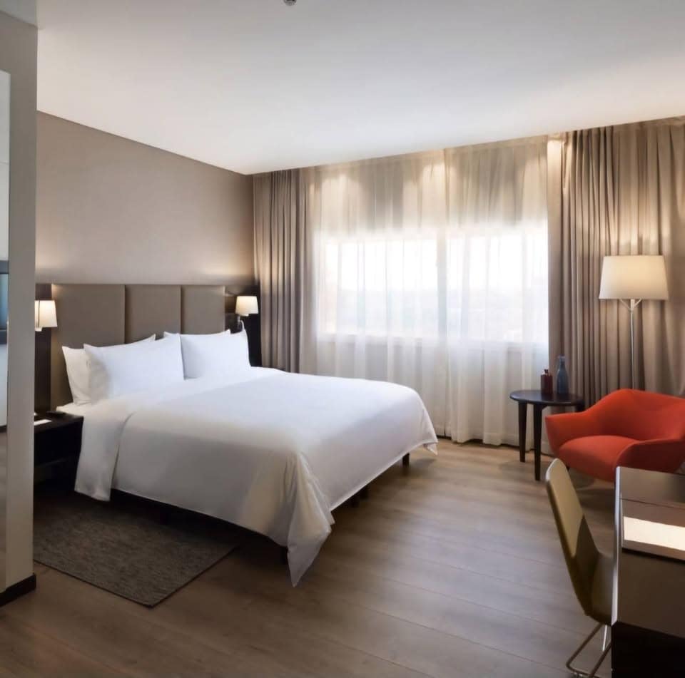 A modern hotel room features a spacious bed dressed in white linens, complemented by soft pillows. Dual bedside lamps provide gentle illumination. A small table and an accent chair are positioned near a large window, allowing natural light to filter in through sheer curtains.