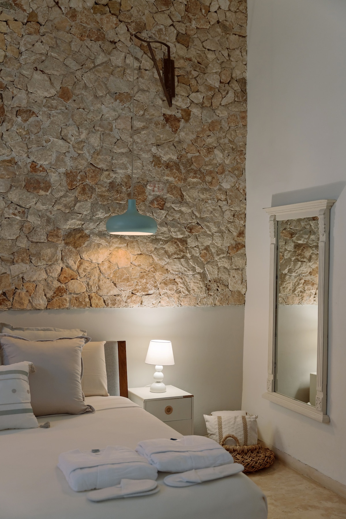 A spacious bedroom features a mampostería stone wall, providing a rustic backdrop. The bed is adorned with elegant linens, and a bedside lamp casts a soft glow. A full-length mirror reflects the room’s calm ambiance, complemented by neatly arranged towels.