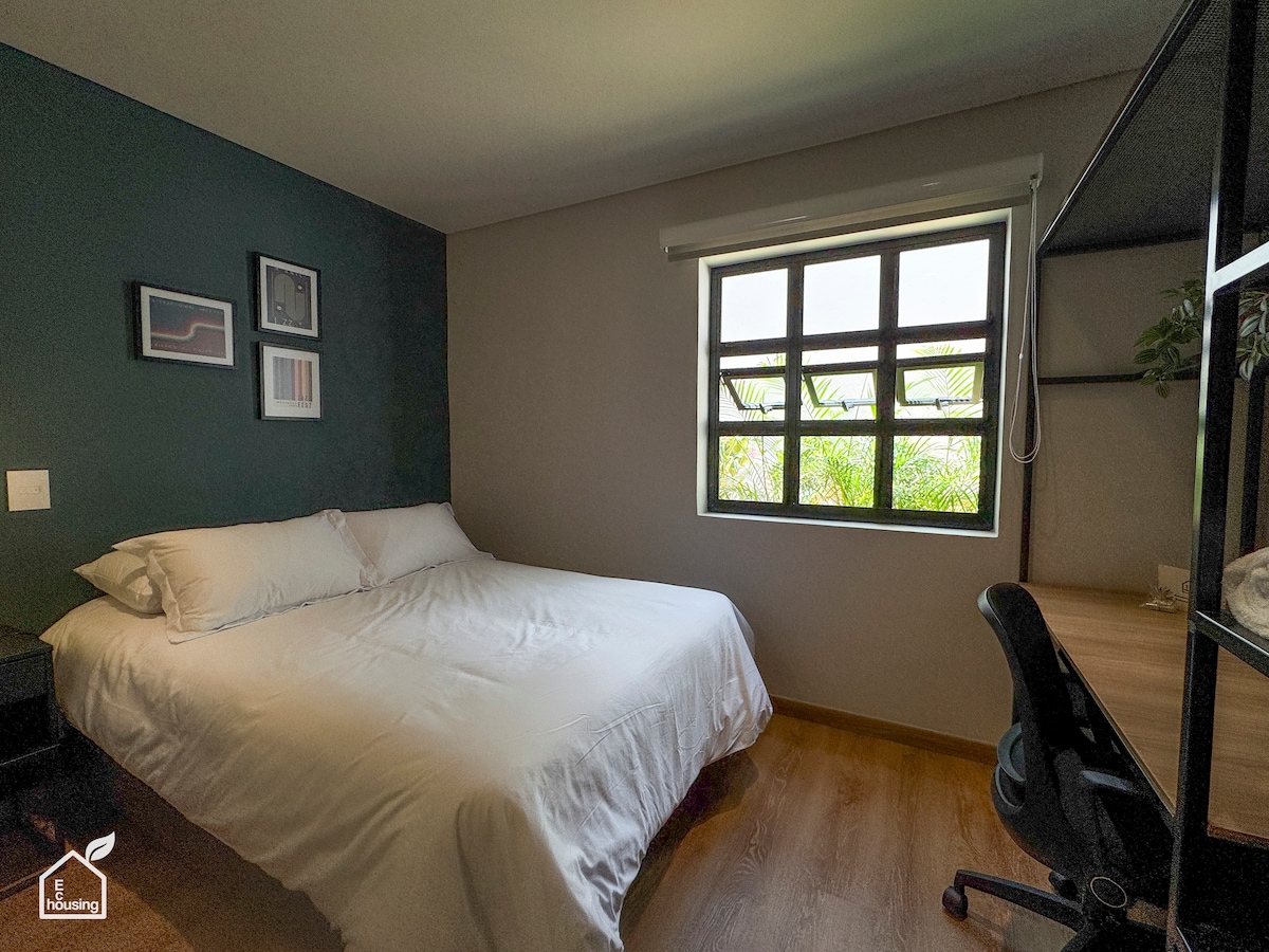A comfortable bedroom features a spacious bed with crisp white linens, complemented by a desk with a chair. Natural light flows in through a window, which offers views of greenery. Artwork is displayed on the walls, adding subtle visual interest.