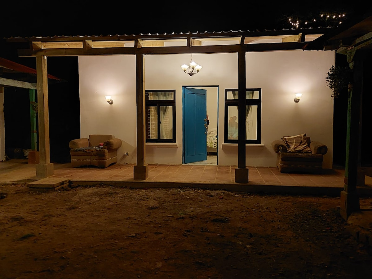 A simple exterior structure is captured at night, featuring a vibrant blue door framed by two wall-mounted lights. Two comfortable seating arrangements occupy the porch, while a warm glow emanates from the lamps. The surrounding area consists of dirt flooring and visible nature.
