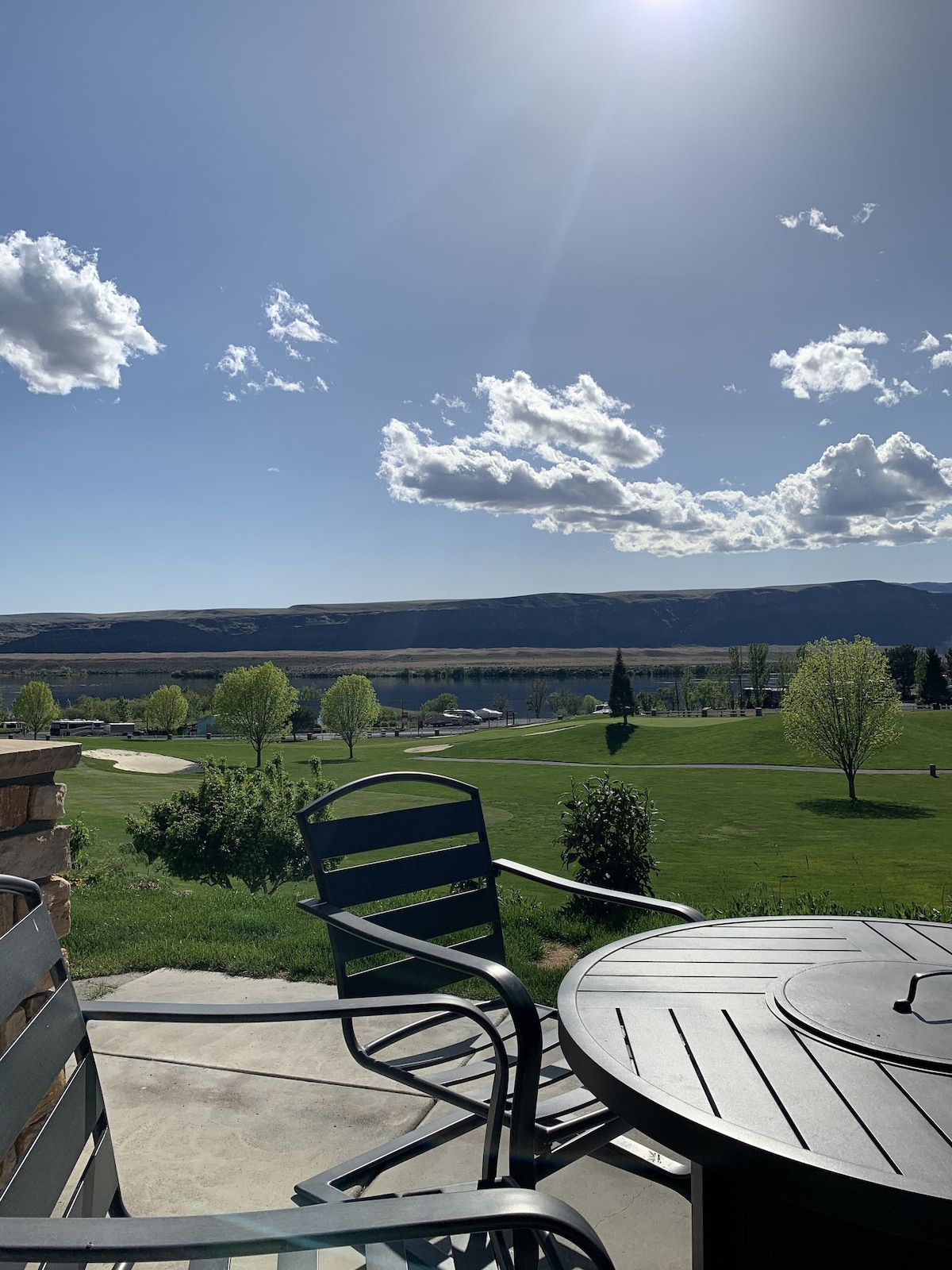 A patio view showcases a serene landscape featuring green lawns and the Columbia River in the distance. Bright sunlight casts shadows over outdoor furniture, where a table is paired with two chairs, offering a relaxing spot to take in the scenery.