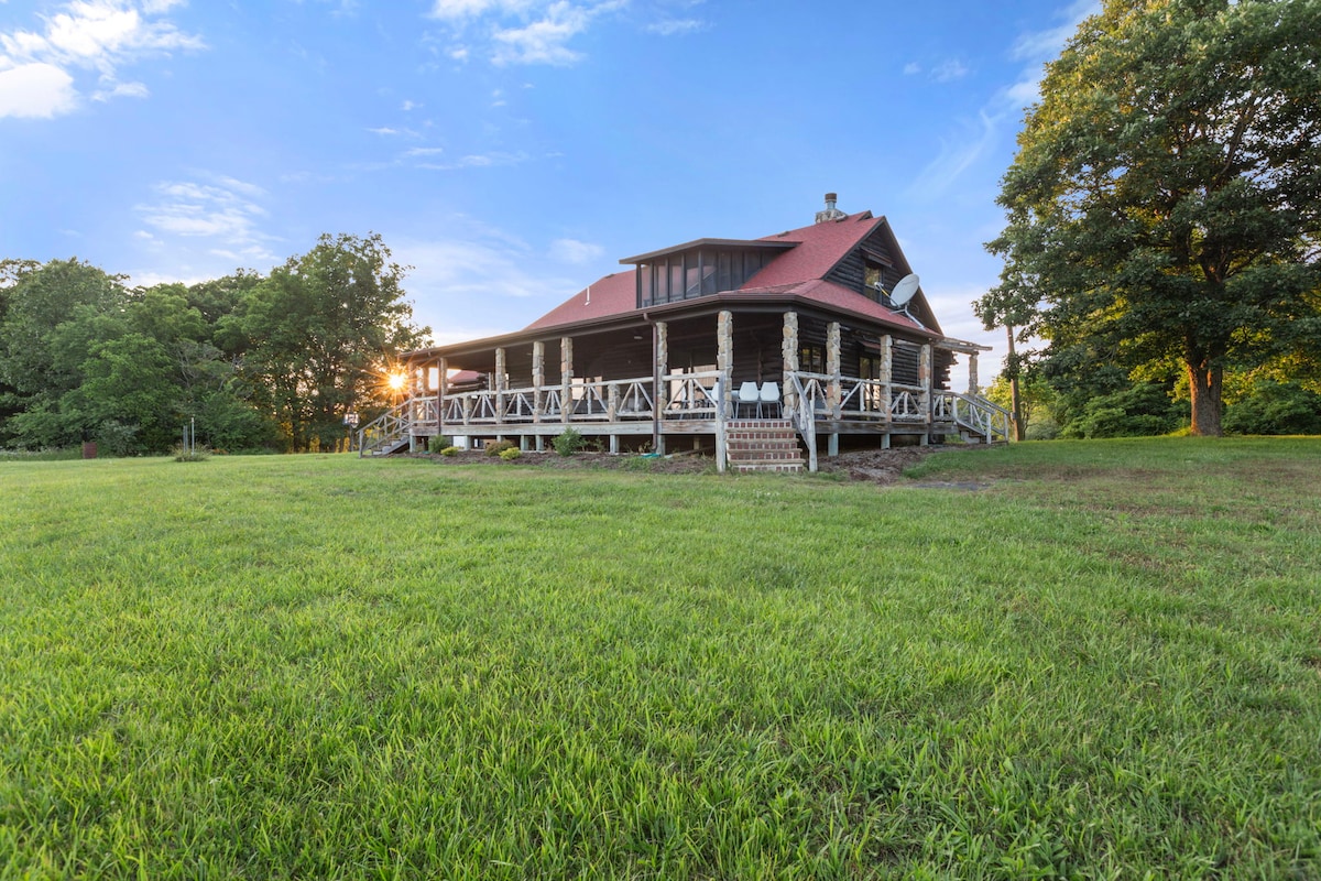 A spacious black log cabin is depicted, elevated on a wrap-around porch overlooking a lush green lawn. Sunlight filters through trees, casting gentle shadows on the grass. The inviting structure features a prominent roof and stone accents, suggesting a harmonious blend with the surrounding nature.