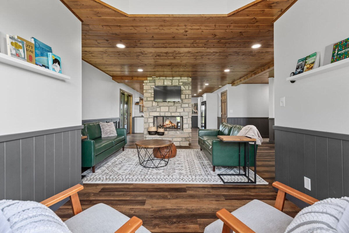 A spacious living area is defined by a stone fireplace, complemented by two green sofas and a wooden coffee table. Natural light fills the room, where a patterned area rug adds texture. Open shelving displays various books, creating a serene and inviting atmosphere.