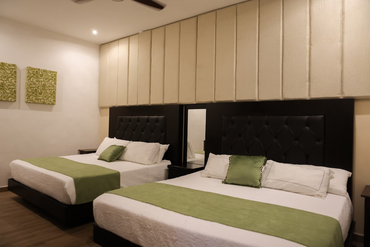 The sleeping area features two spacious beds with light green accents and decorative pillows. A wall-mounted mirror reflects the stylish headboards, while soft wall covers create a comfortable environment. Two pieces of wall art in shades of green add a subtle touch of decor.