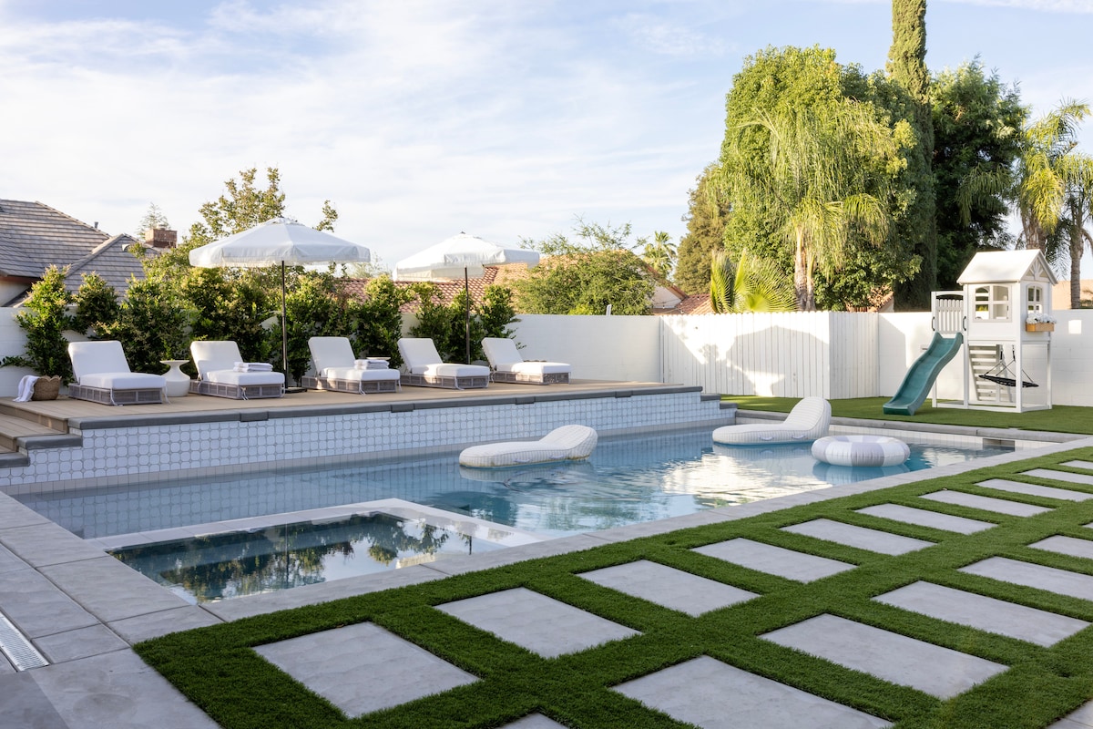 An outdoor space features a kidney-shaped pool surrounded by lounge chairs. A small slide and playhouse are visible on the lawn. Umbrellas provide shade for relaxation, while neatly arranged pavers lead across green grass, enhancing the recreational area.