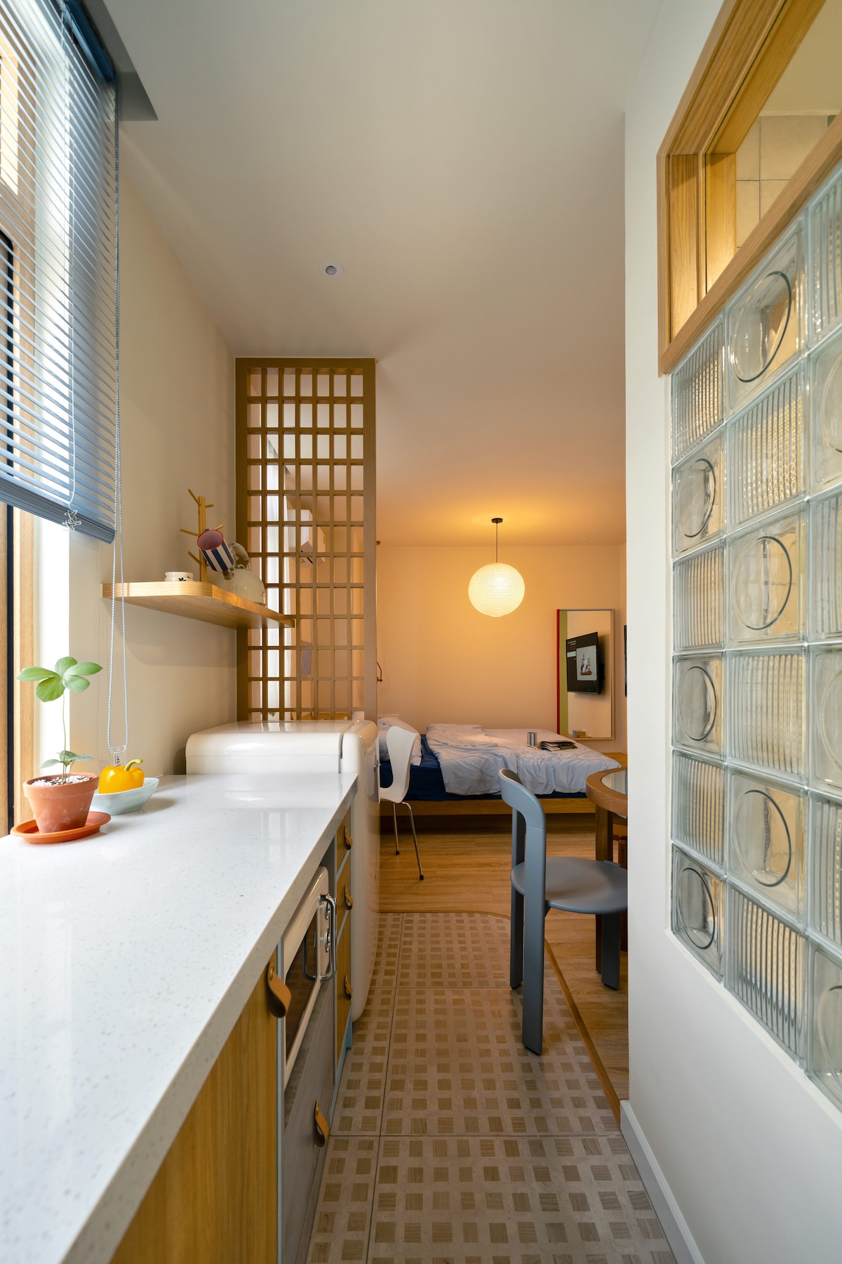 The image displays a cozy kitchenette featuring a white countertop and wooden cabinetry. A small potted plant adds a touch of greenery. A warm light illuminates the adjacent space, which includes a glimpse of a bedroom with a spacious bed, framed in soft colors and minimal decor.