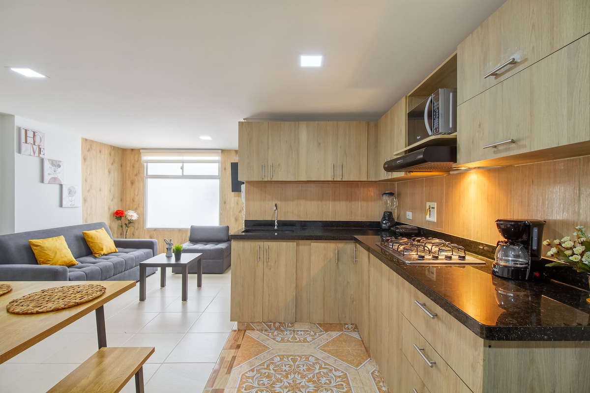 A modern kitchen area features wooden cabinetry and a dark countertop, equipped with a gas stove and coffee maker. Adjacent is a cozy seating area with a sofa and bright cushions, accompanied by a small table holding a vase of fresh flowers.