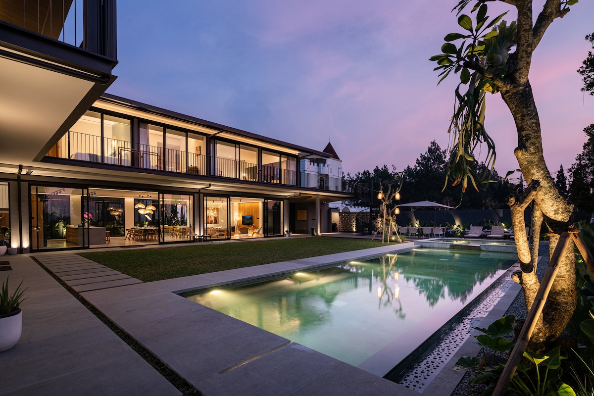 A modern villa is showcased against a twilight sky, featuring expansive glass walls and a private swimming pool reflecting soft hues. Lush greenery surrounds the outdoor space, while cozy seating areas invite relaxation by the water. Natural elements accentuate the contemporary design.