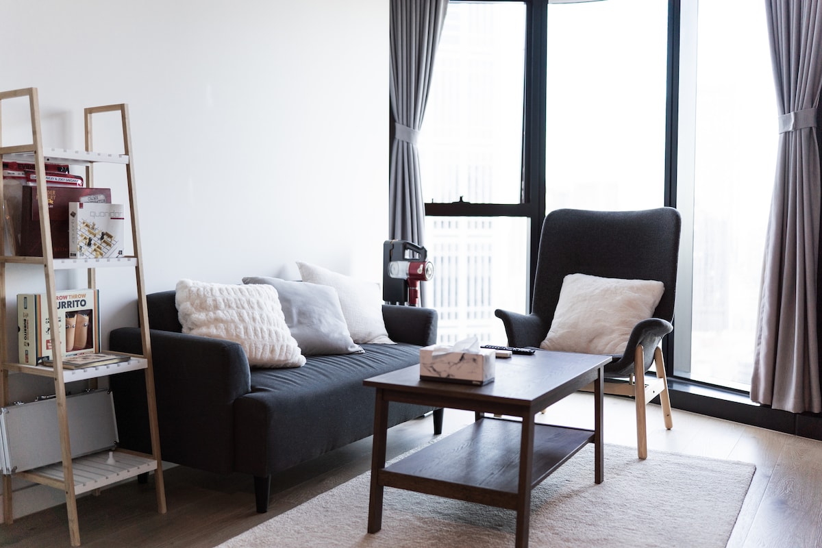 A modern living room is showcased with a sleek dark sofa and an armchair, complemented by a wooden coffee table at its center. A bookshelf filled with magazines is positioned to one side, while large windows allow natural light to fill the space.