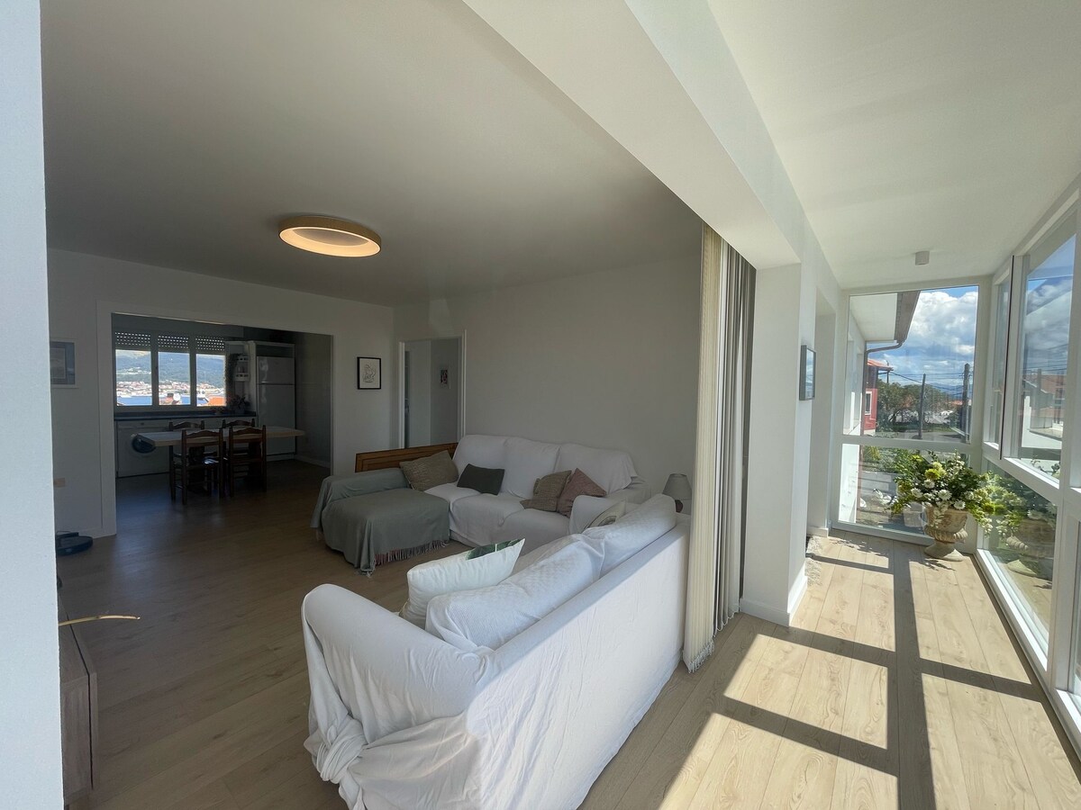 A bright and spacious living area showcases a white sofa and a wooden chair. Natural light floods through large glass windows. A dining space with a wooden table and chairs is visible in the background. A serene terrace helps to connect indoor and outdoor living.