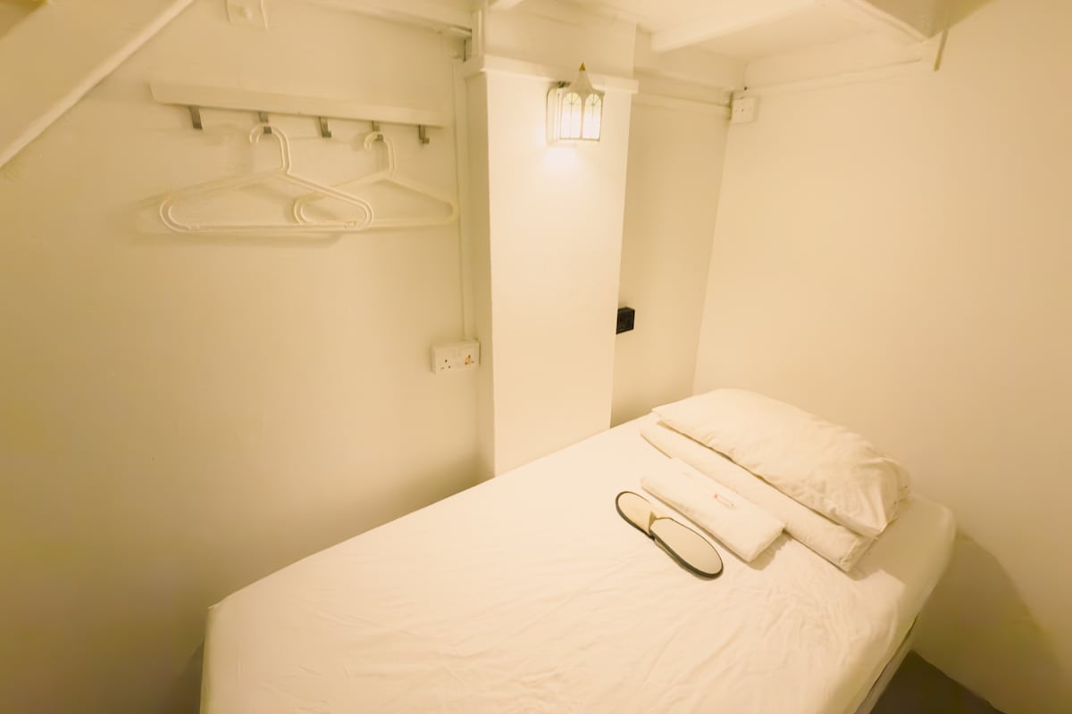 A compact cabin pod features a single bed with fresh linens and a pillow. A small bedside table holds a towel and dental kit. The wall is adorned with a lantern, while hooks are positioned above for hanging items.