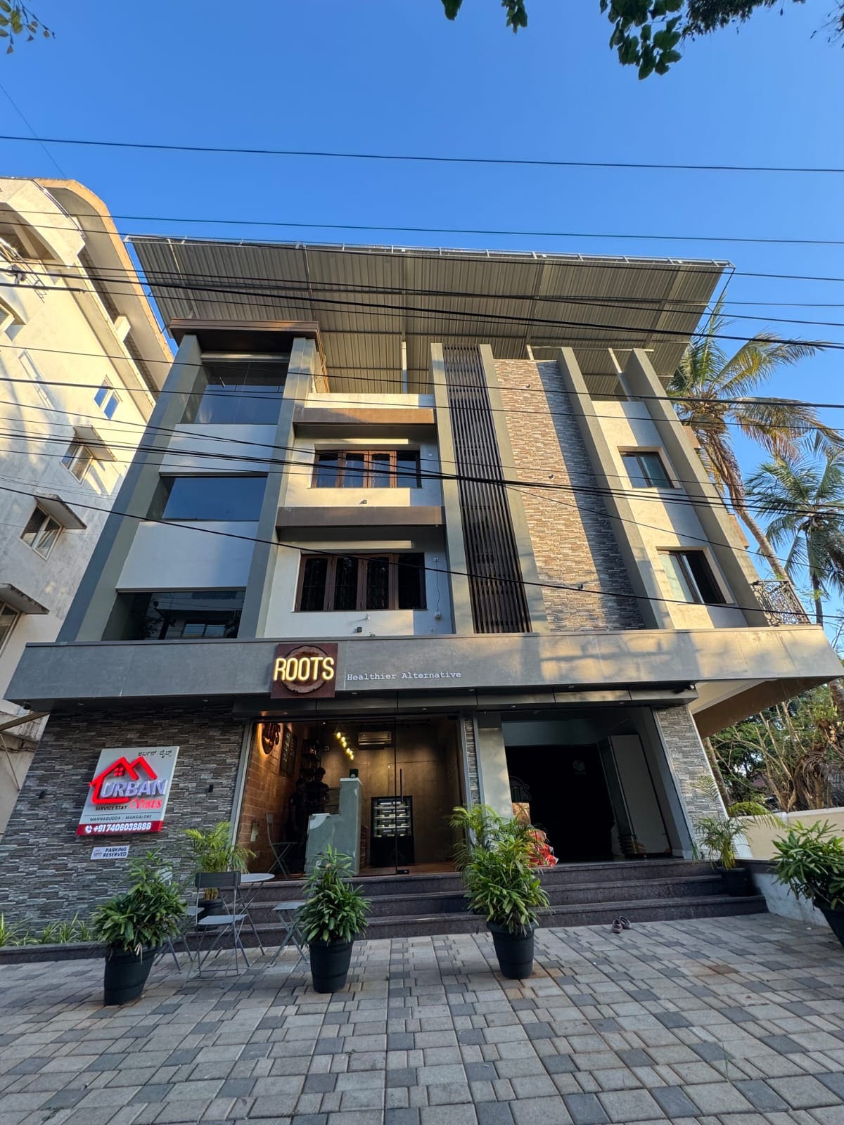 The exterior of a modern three-story building is showcased, featuring a combination of stone and concrete materials. The entrance is marked by a prominent sign stating 'ROOTS,' with potted plants flanking the steps. Large windows allow natural light into the structure, and palm trees are visible in the background.