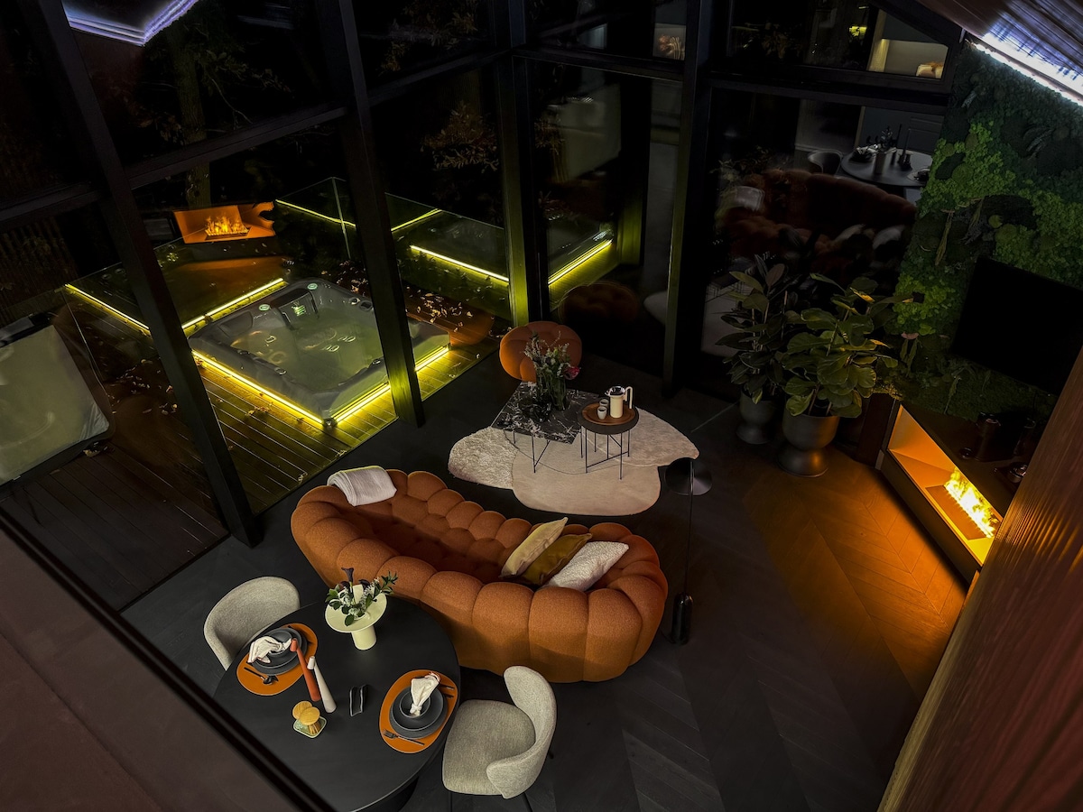 A stylish living area features a curved orange sofa, complemented by a round coffee table and two dining chairs. Large glass walls provide a view of an outdoor space illuminated by soft lighting, enhancing the tranquil atmosphere of the cabin.