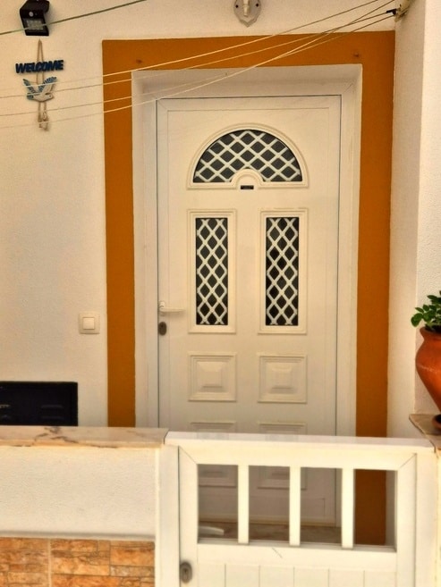 A white front door with a decorative lattice design is framed by an orange wall. A welcome sign hangs above the door, while a planter with greenery is positioned to the right, enhancing the entrance's inviting appearance.