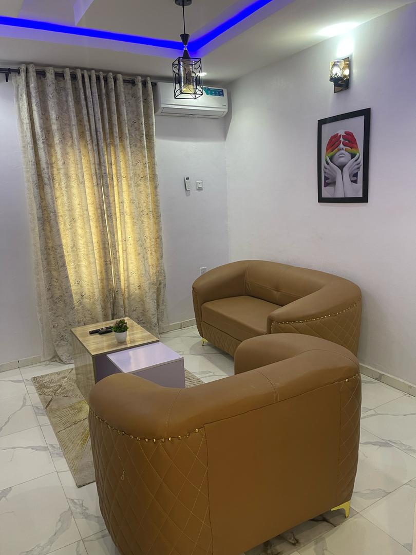 A cozy seating area is displayed with two rounded armchairs in a warm, tan color, positioned around a small white table. A light grey rug lies beneath them, and soft curtains filter warm light from a nearby window. Artwork adorns the wall, adding a touch of style.