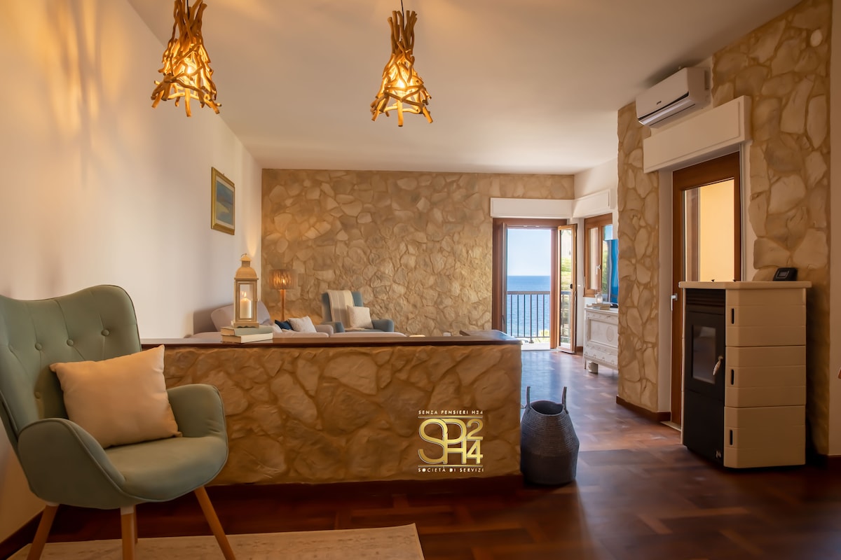 A spacious living area features stone walls and wooden flooring. Two large armchairs and a sofa are arranged for seating, with large glass doors providing views of the sea. Natural light is diffused through sheer curtains, creating a calm and airy environment.
