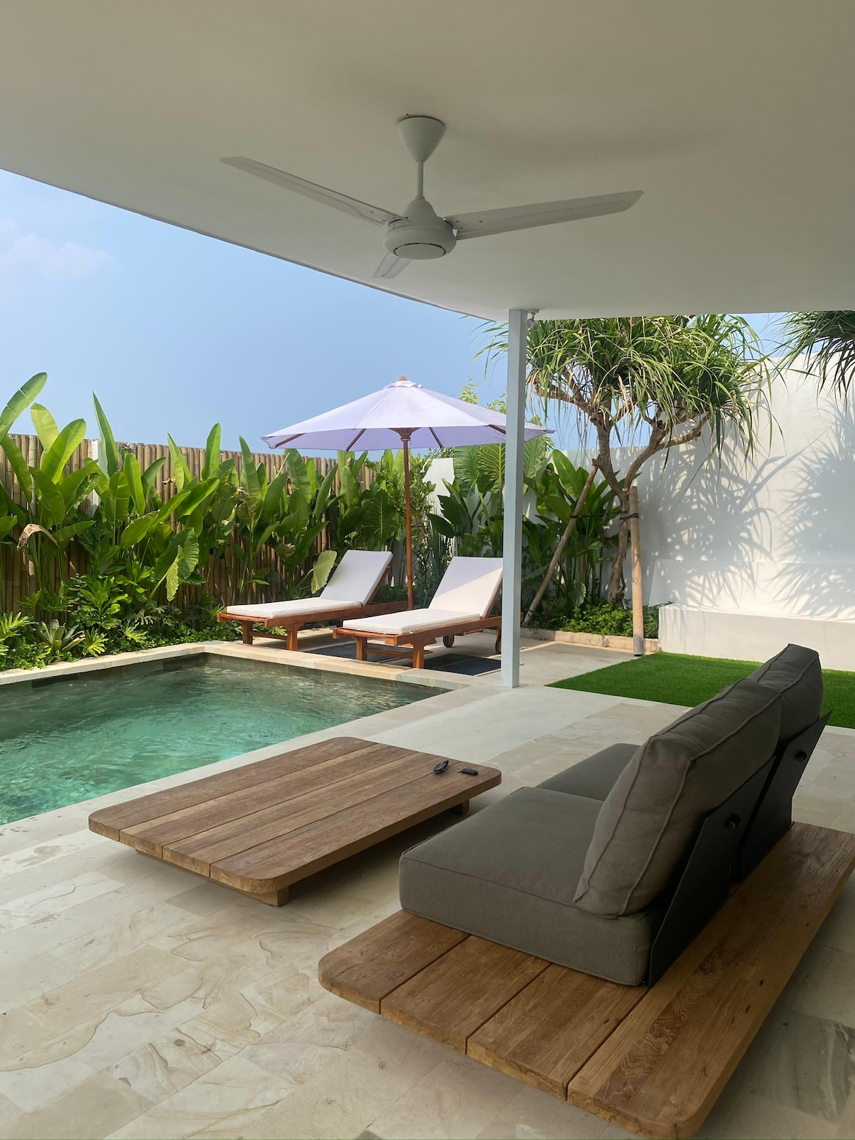 A relaxing outdoor seating area features a gray cushioned sofa and a wooden coffee table. Two sun loungers are positioned beside a small pool, shaded by a white umbrella. Lush greenery creates a private ambiance, complementing the clean lines of the villa.