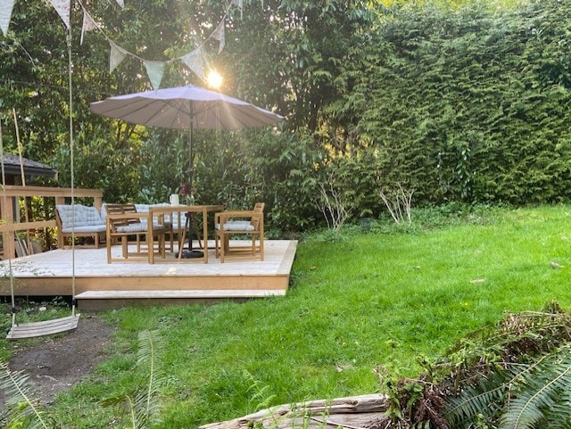 An outdoor deck is surrounded by lush greenery, featuring a large umbrella providing shade over a wooden table and chairs. A swing hangs nearby, and sunlight filters through the trees, creating a serene atmosphere in the garden space.