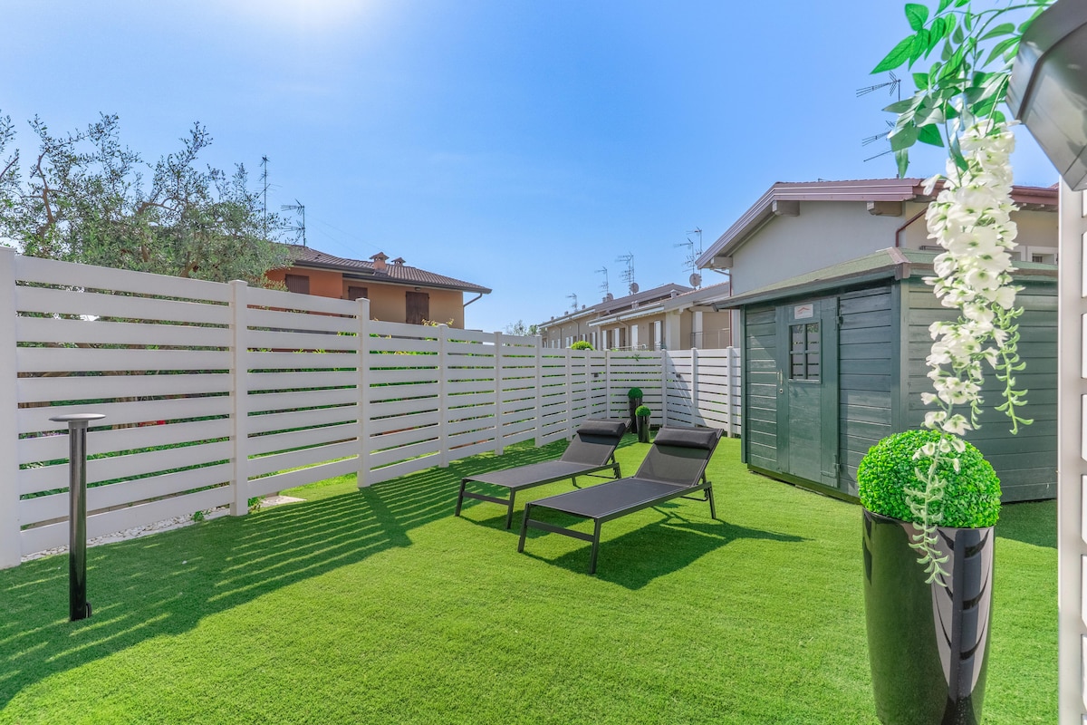 A private garden space features a well-maintained lawn, two lounge chairs for relaxation, and a small storage shed. A tall decorative planter adds a touch of greenery, while a white fence provides privacy. The area is bathed in natural light under a clear blue sky.