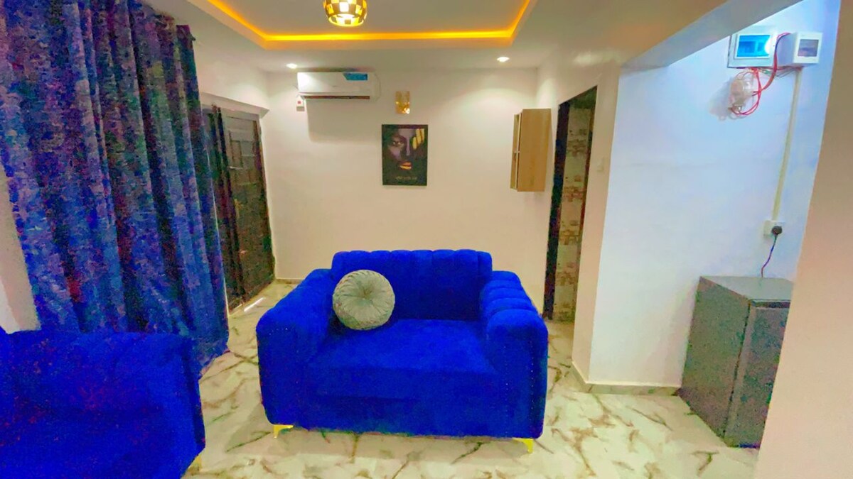 A cozy seating area features two plush blue sofas facing each other. A gray cushion rests on one sofa. The space is illuminated by a warm ceiling light, while dark curtains frame a window. A small, functional side table is positioned against the wall.