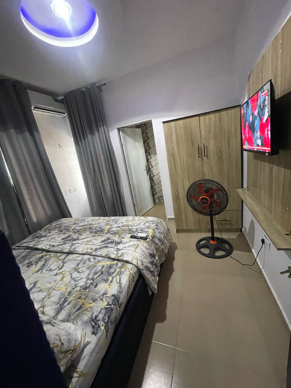 A cozy bedroom features a comfortable bed topped with a decorative blanket. Medium-toned curtains frame the window, and a wall-mounted television offers entertainment. A standing fan provides airflow, while a wooden cabinet is positioned against the wall, creating a functional and relaxing environment.