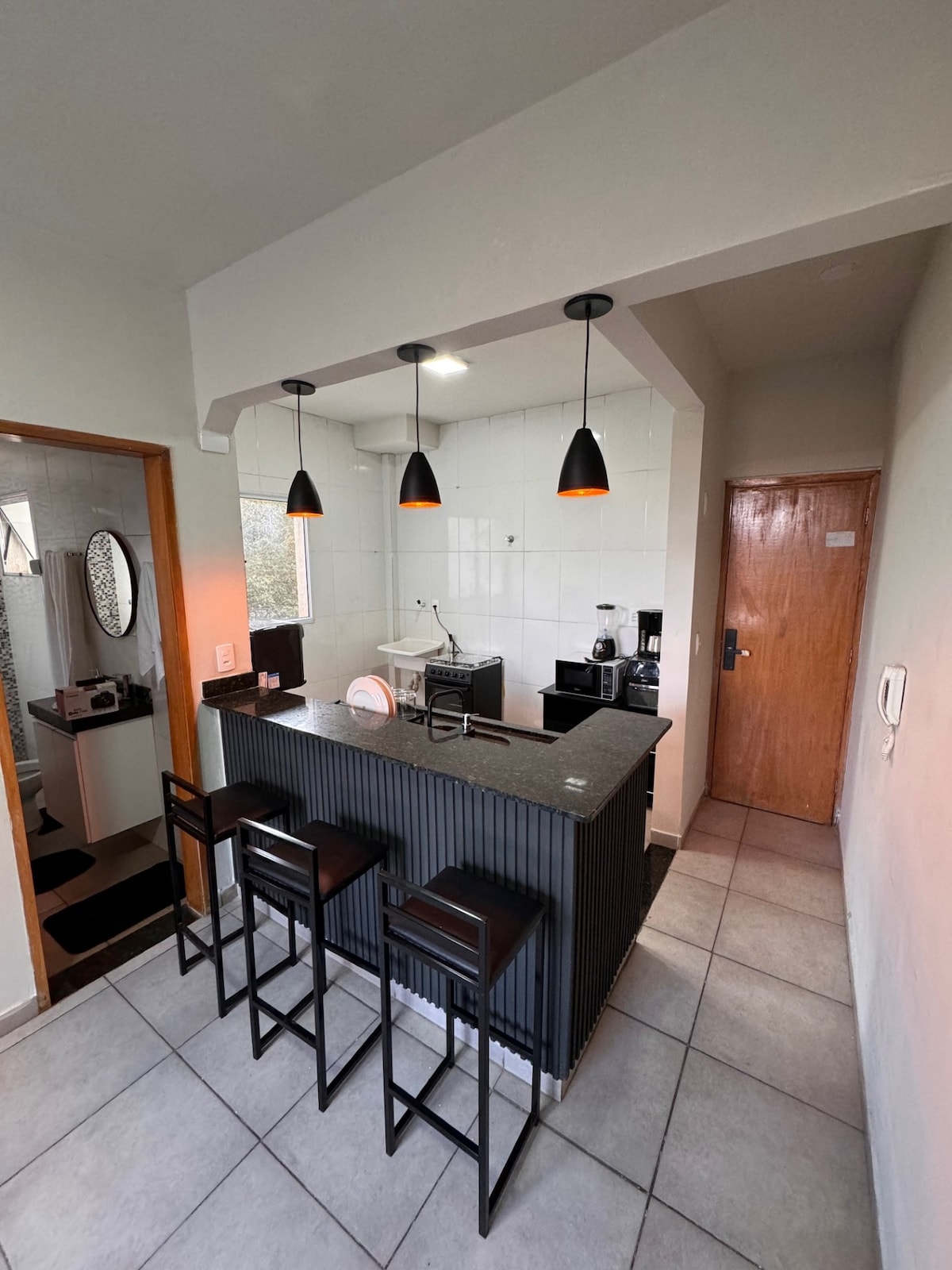 A compact kitchen area is visible, featuring a granite countertop with three bar stools positioned alongside. Pendant lights hang above the counter, illuminating modern appliances, including a coffee maker and kettle. The tiled floor and light-colored walls contribute to the clean aesthetic.
