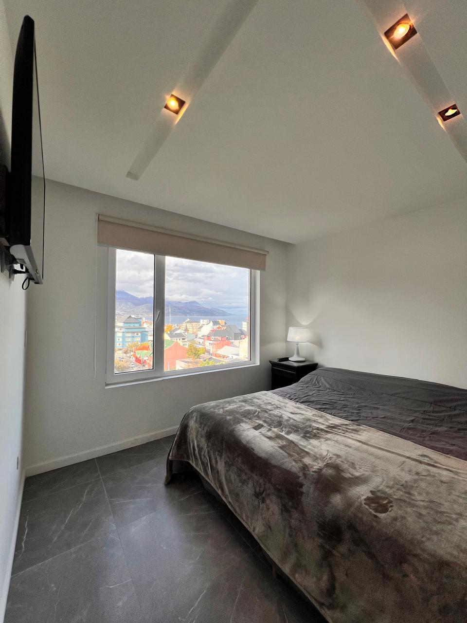 A bright bedroom features a spacious bed covered with a soft gray blanket. A bedside table with a lamp is positioned next to the bed. A large window offers a view of the outdoors, while recessed lighting enhances the modern design.