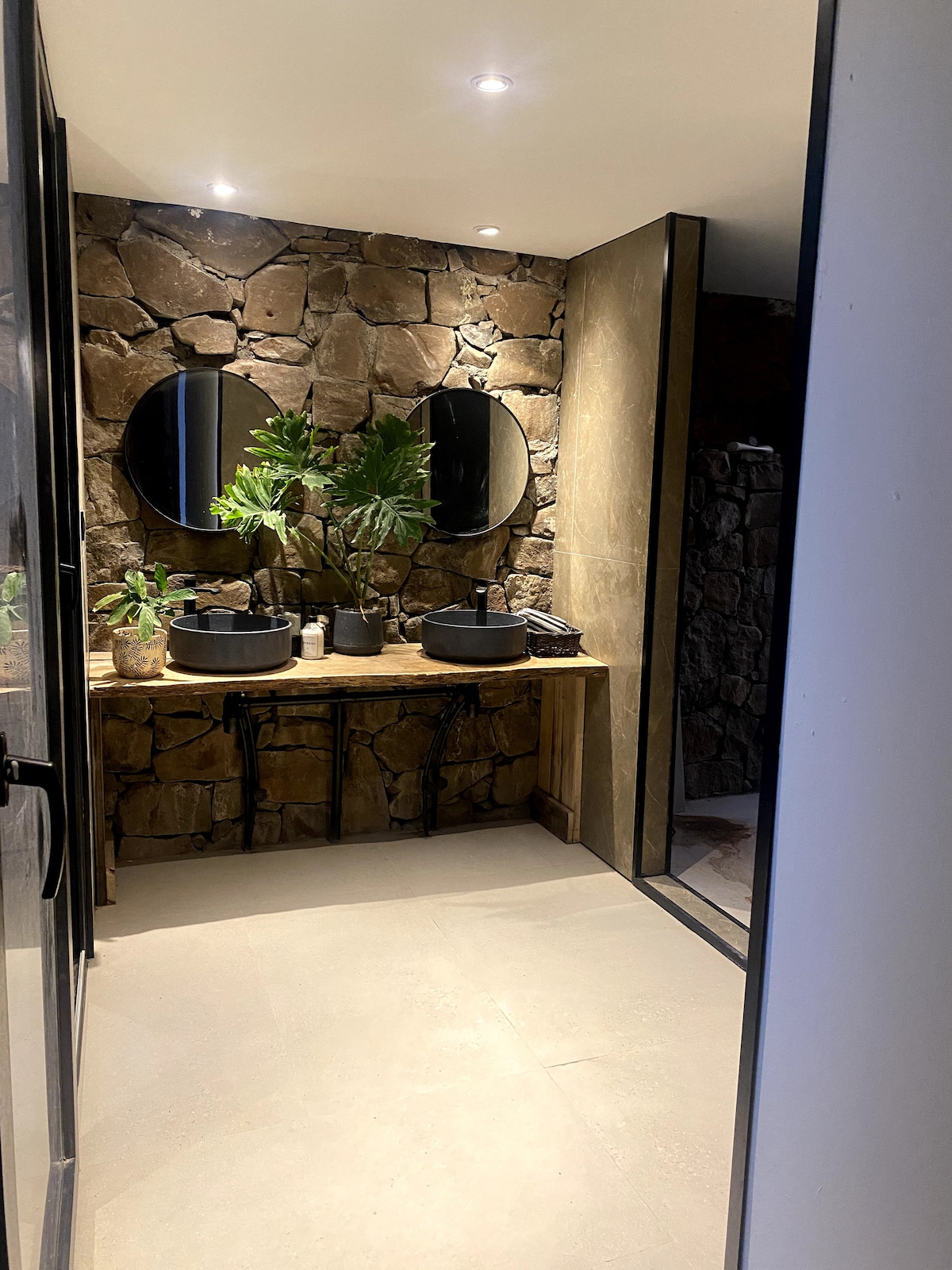 The bathroom features a natural stone wall and a wooden vanity with two round mirrors above. Two circular sinks are placed on the vanity, complemented by small potted plants. Soft lighting enhances the warm tones of the materials used in the space.