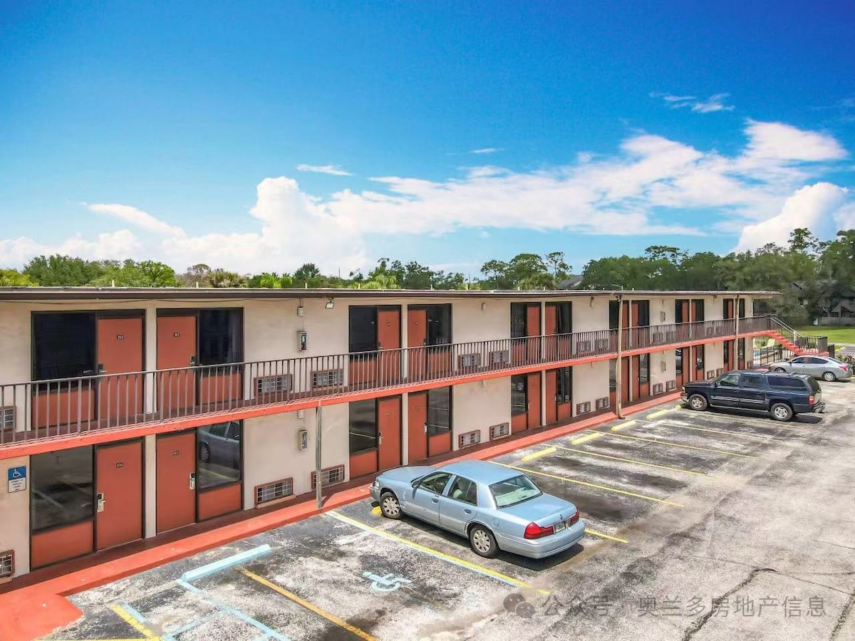 An exterior view of a two-story motel showcases a series of rooms with orange accents and black railings. Several parked cars are visible in the foreground, with a clear blue sky and green trees in the background.