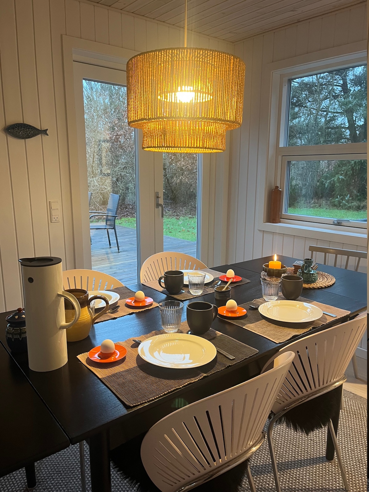A dining area is presented with a dark table set for several guests. Plates, cups, and utensils are arranged neatly alongside decorative candles. A pendant light hangs overhead, while large windows invite natural light and offer a view of the outdoors.
