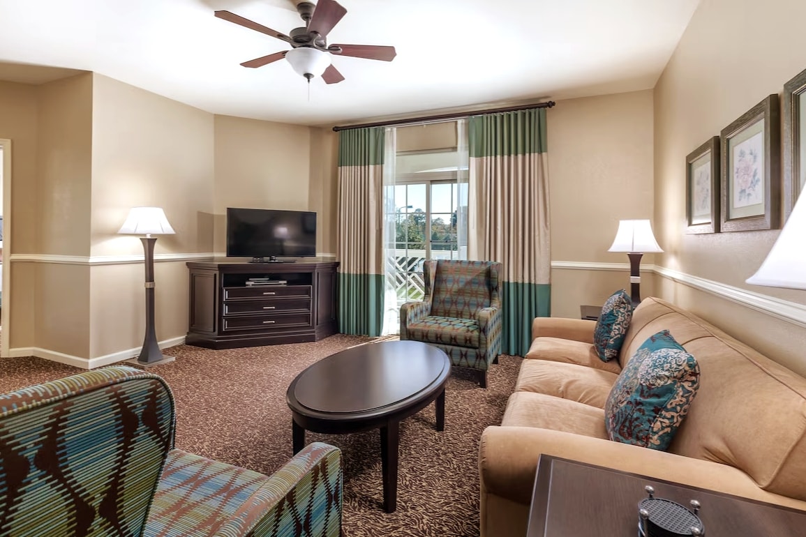 A welcoming living area is presented with a comfortable sofa and two patterned chairs. A dark coffee table centers the space, with a flat-screen TV placed on a modern console. Two lamps provide soft lighting, while large windows with patio access offer natural light.