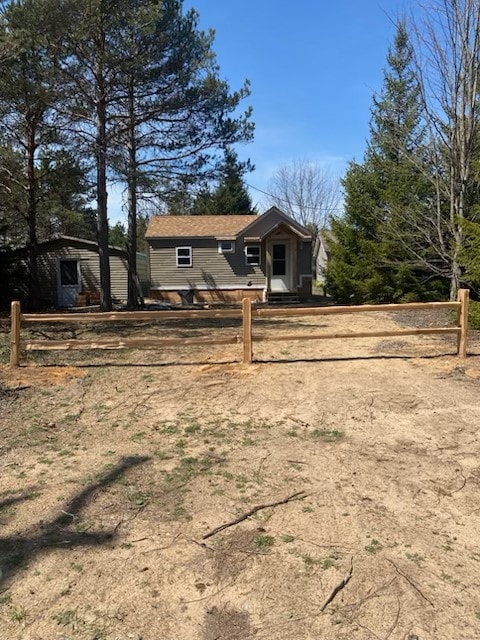 A modest cottage is set amidst tall trees, featuring a welcoming front porch. A natural dirt path leads to a wooden fence that encloses the property. Sunlight filters through the branches, highlighting the simplicity of the outdoor space.
