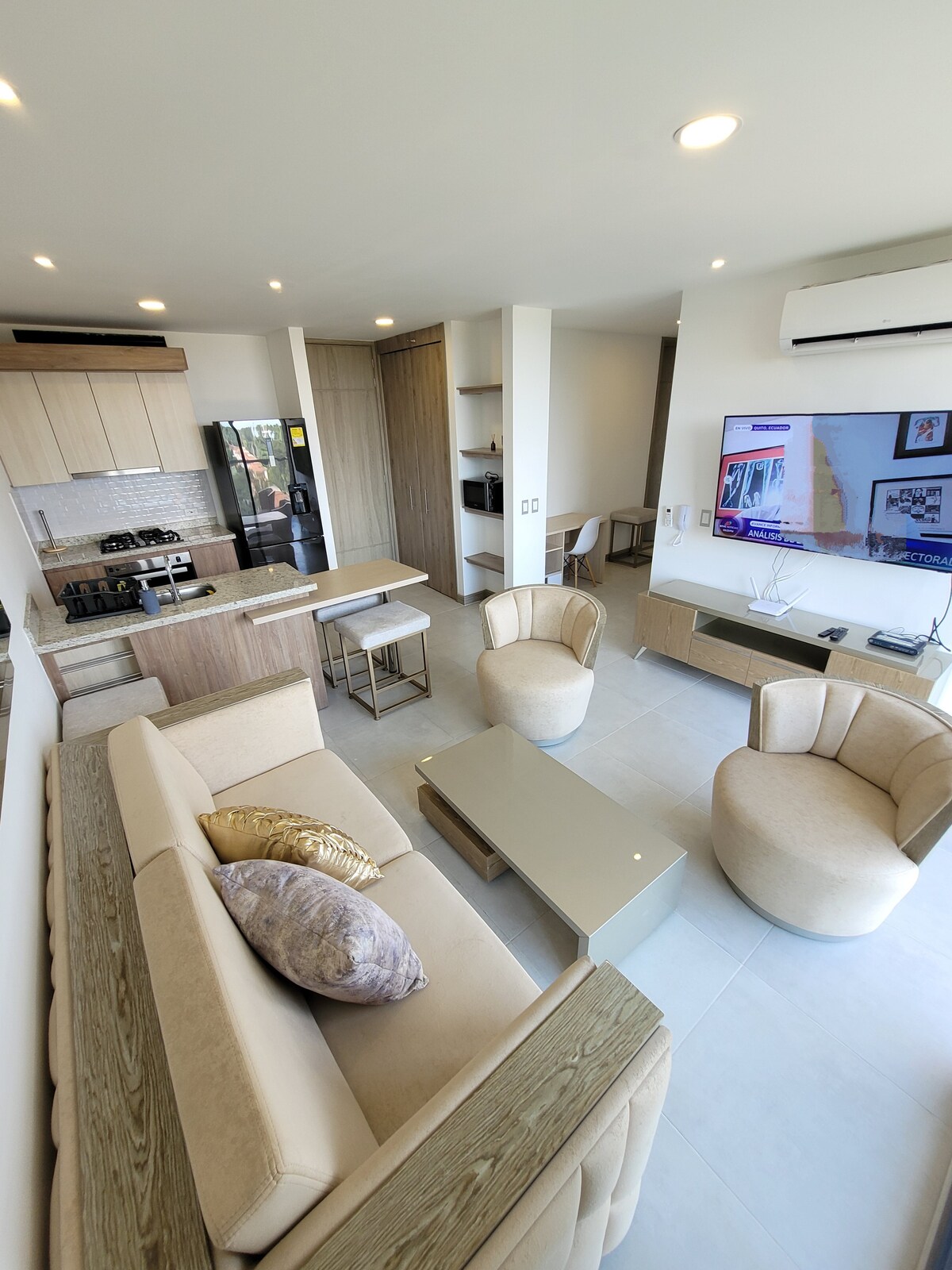 The living area features a light-colored sofa with decorative cushions, paired with two modern armchairs. A sleek coffee table sits at the center, while a wall-mounted Smart TV is visible. Natural light enters through the large window, illuminating the open design.