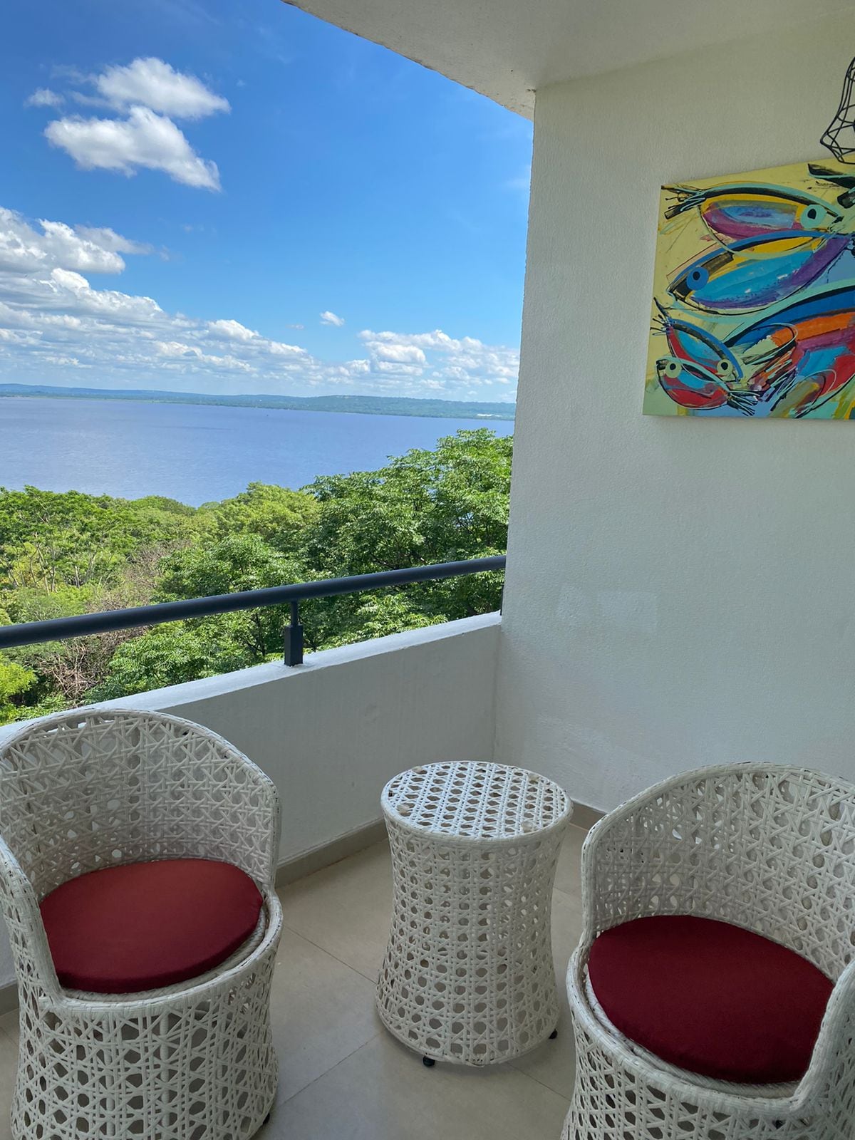 A charming balcony features two white wicker chairs with red cushions and a small round table. In the background, expansive views of greenery and a shimmering body of water are framed by a clear blue sky adorned with fluffy clouds.
