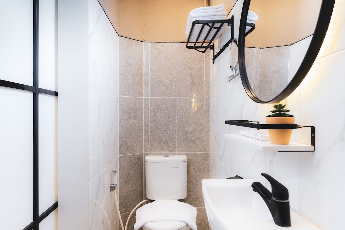 A modern bathroom features sleek tile walls and a stylish round mirror. A compact toilet and a minimalist sink are positioned below the mirror, while a floating shelf holds neatly arranged towels and a small potted plant. Soft lighting enhances the contemporary design.