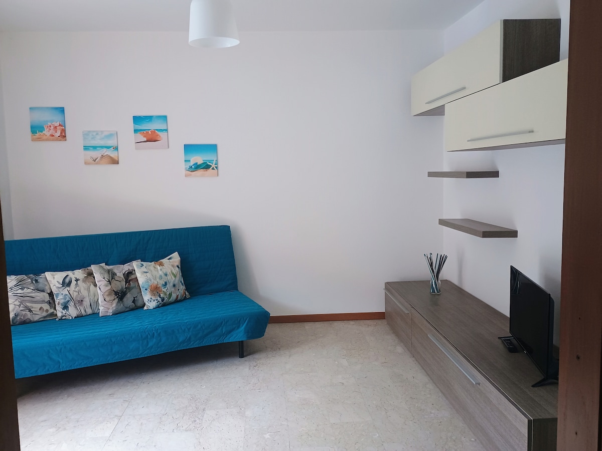 A bright living area features a blue sofa bed adorned with patterned cushions. Above, four beach-themed wall art pieces add a touch of color. A modern entertainment unit sits against the wall, complemented by a small black television and a minimalist shelf.