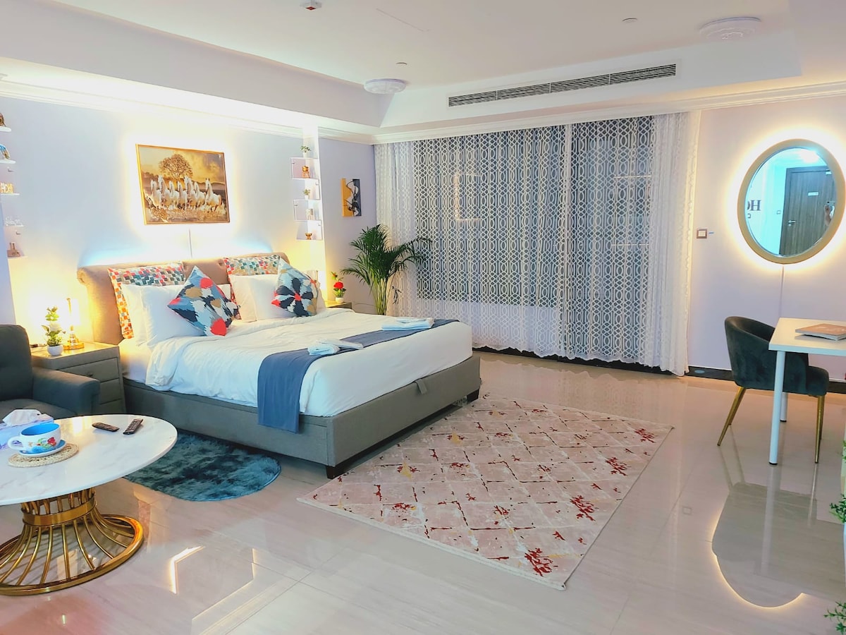 A modern studio layout is observed featuring a spacious bed adorned with decorative pillows and a plush blanket. A light-colored rug complements the marble floor beneath a round mirror. A small dining table and a comfortable seating area complete the inviting room.
