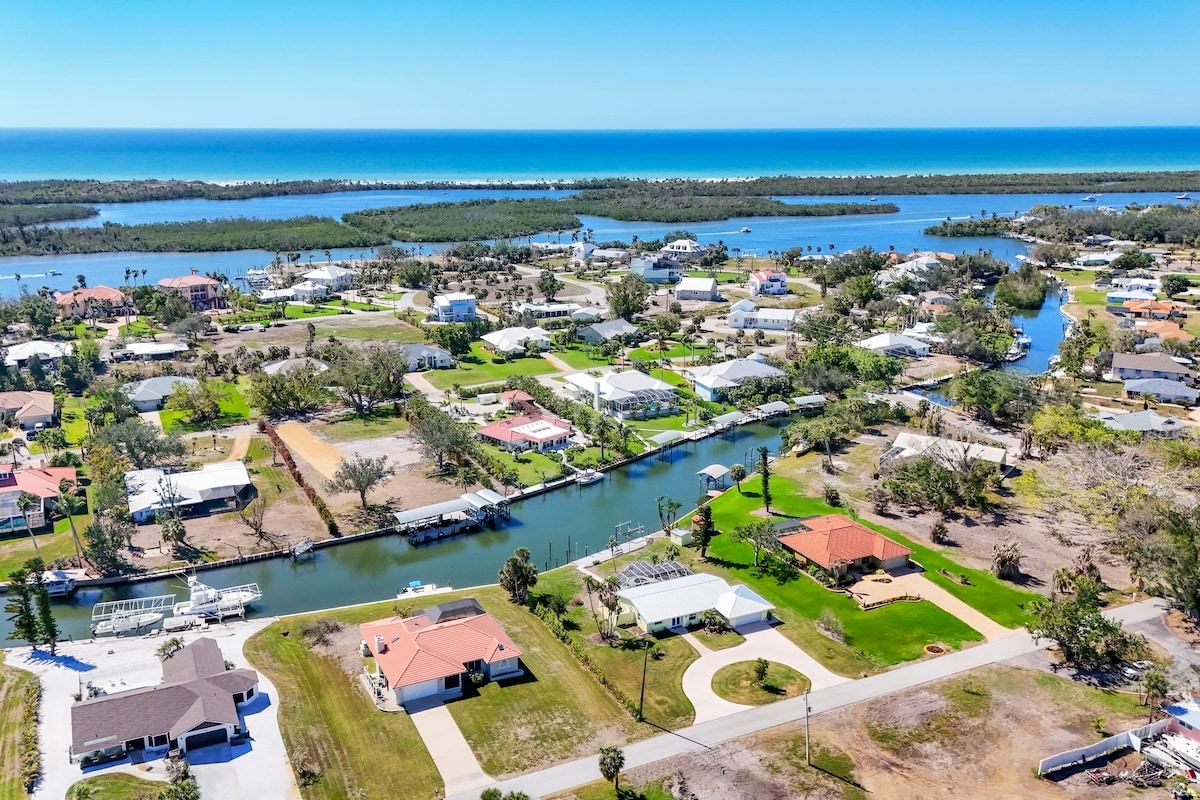 An aerial view showcases the serene neighborhood with waterways and the Gulf of America visible in the distance. Houses are nestled along the coast, surrounded by lush greenery and palm trees, creating a tranquil atmosphere. The layout highlights convenient access to boating and nearby beaches.