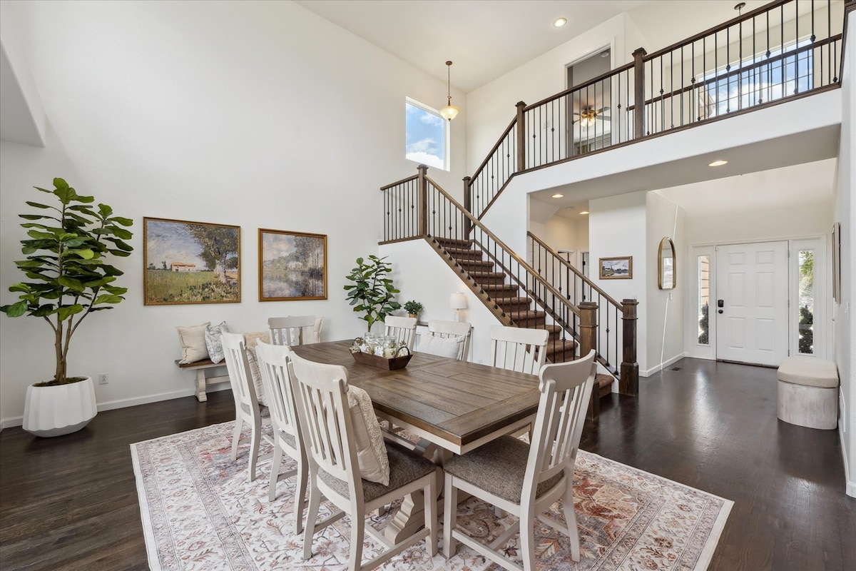 A spacious dining area is featured, with a wooden table surrounded by eight light-colored chairs. Large windows provide natural light, illuminating the space. A staircase leads to the upper level, while decorative plants and artwork enhance the inviting atmosphere.
