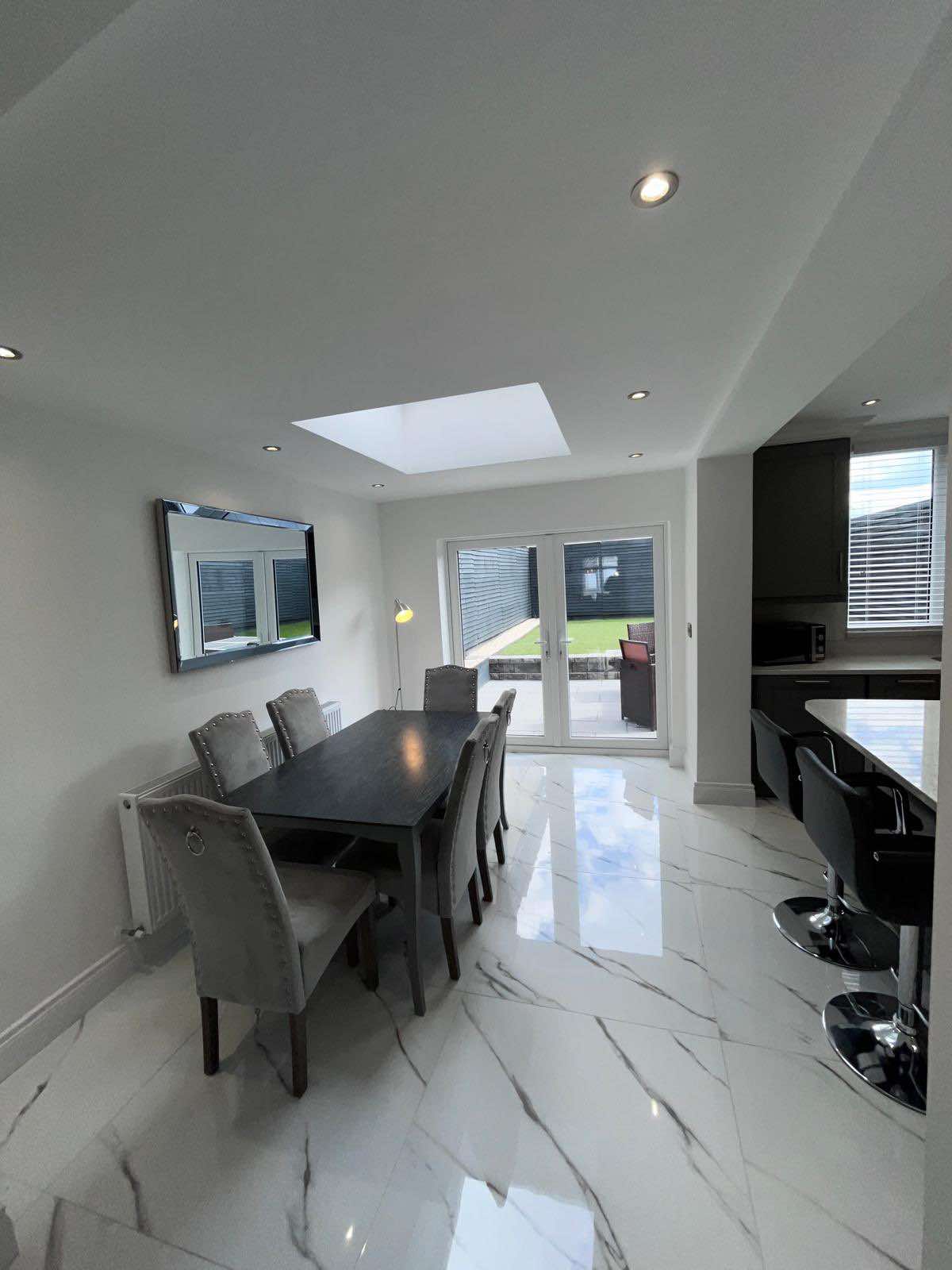 A modern dining area is shown with a sleek table surrounded by six upholstered chairs. Natural light enters through a skylight, complementing the marble tiled floor. Glass doors lead to the outdoor patio, enhancing the open and airy feel of the space.