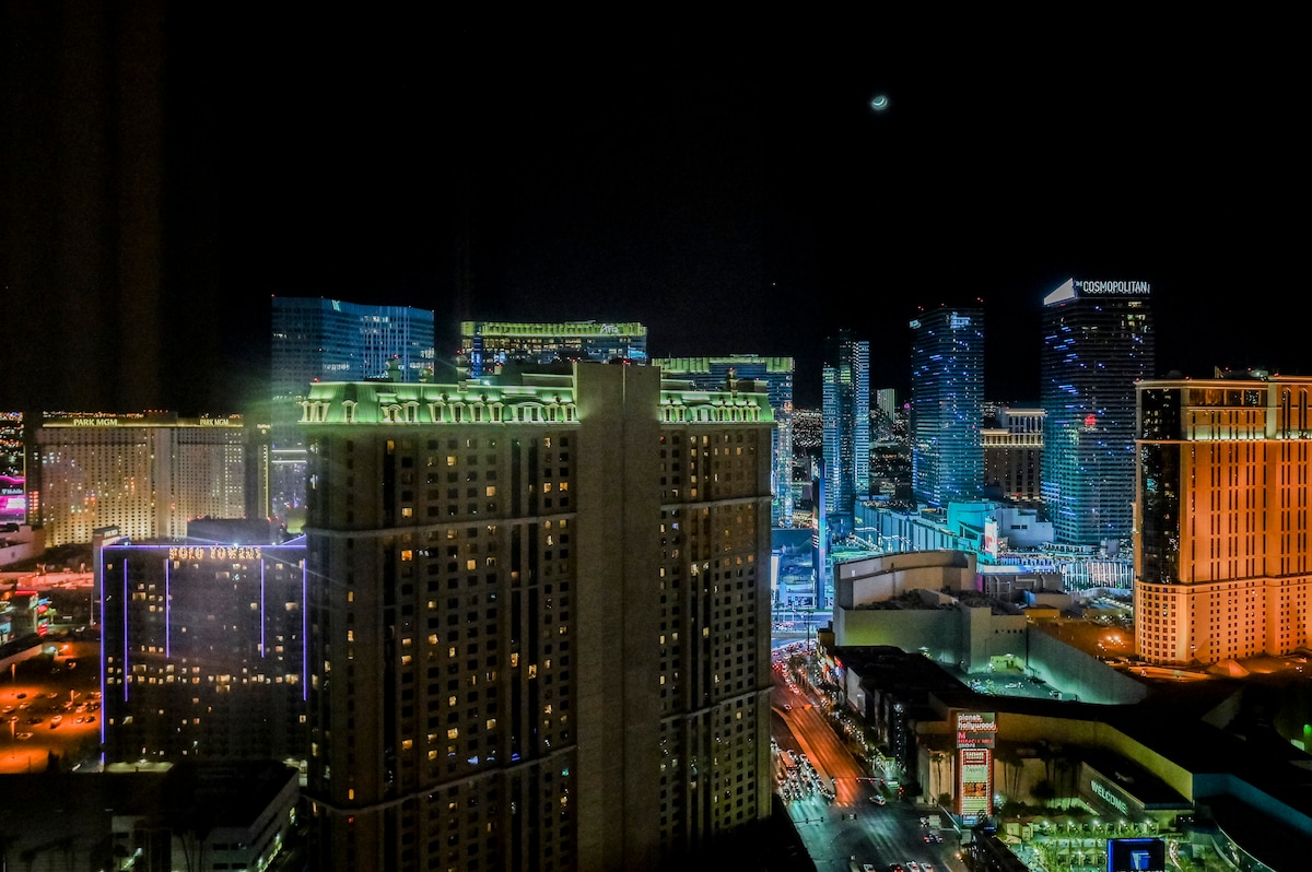The vibrant Las Vegas Strip is illuminated at night, with high-rise hotels and casinos featuring colorful lights. A distant view of the bustling cityscape reveals diverse architectural styles. Streets below showcase movement, hinting at the lively atmosphere of the area.