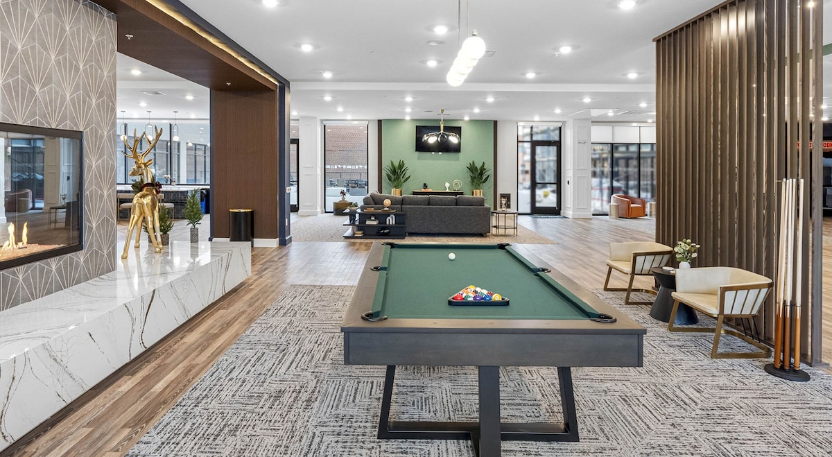 A spacious common area features a billiards table at the center, surrounded by stylish seating arrangements. Natural light filters through large windows, illuminating modern decor elements, including a decorative feature wall and seating options, creating an inviting environment for relaxation and socializing.