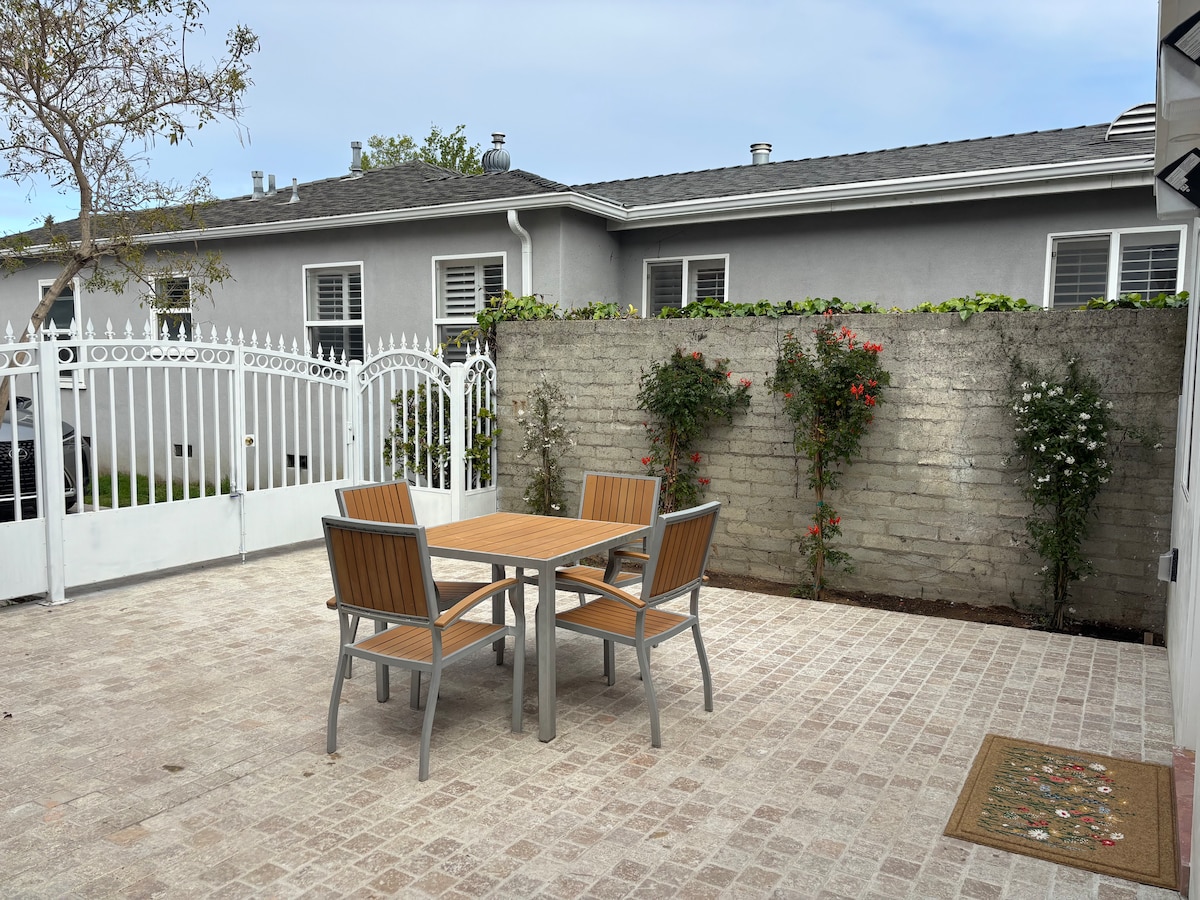 A private outdoor patio features a wooden table surrounded by four matching chairs. A textured stone wall adorned with climbing plants offers a natural touch. The space is paved with light-colored tiles, providing a serene area for relaxation.