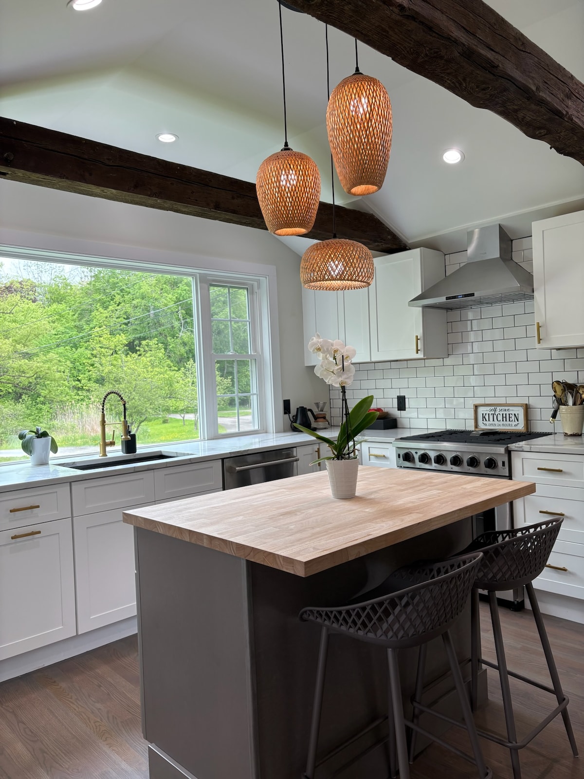 A well-equipped kitchen features modern appliances and a spacious central island with bar stools. Natural light floods the room through a large window, showcasing views of the greenery outside. Wooden beams add a touch of character, while pendant lights enhance the warm ambiance.