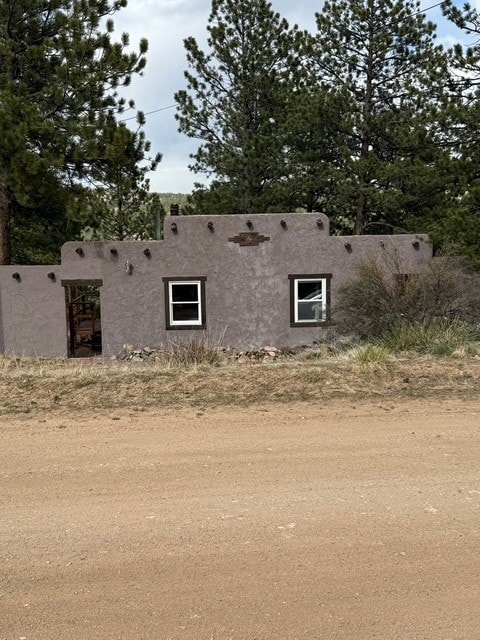 A charming cabin structure is set against a backdrop of tall pine trees. The exterior features a smooth, muted facade with two windows symmetrically placed, allowing natural light to enter. The surrounding area is characterized by dry grasses and earthy tones, creating a tranquil rural setting.