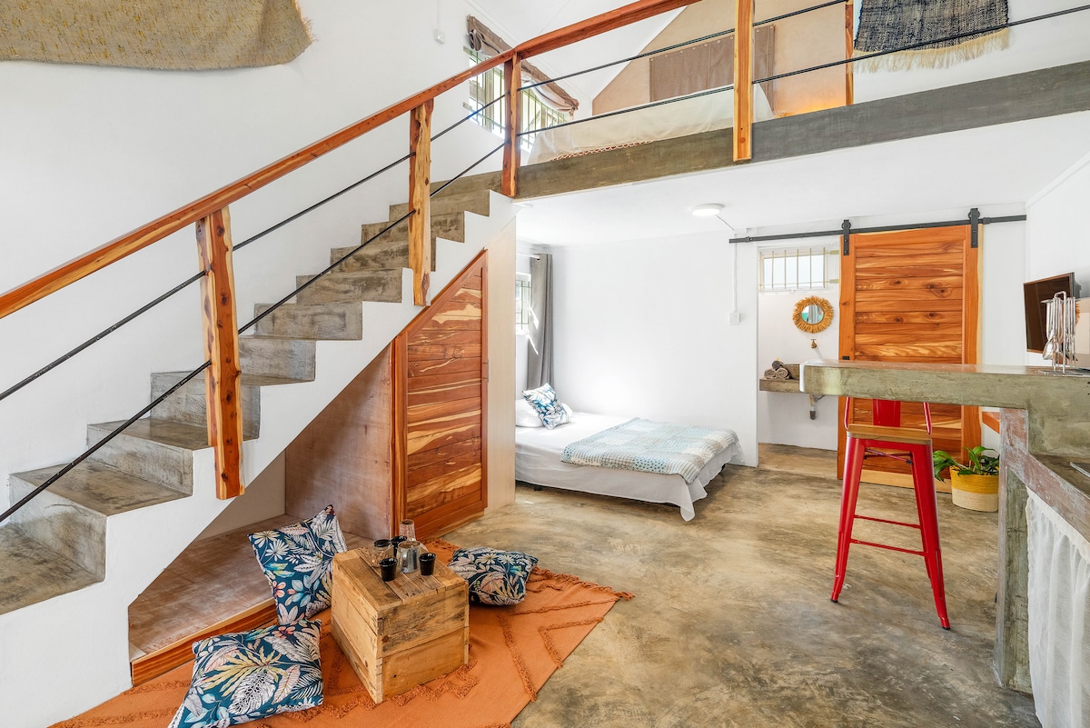 The interior space features a two-level layout with wooden stairs leading to a mezzanine sleeping area. Natural materials and a warm color palette are present, including a cozy seating area on the textured rug and a small table with vibrant accents in the foreground.