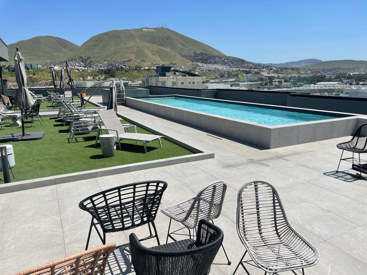 A rooftop area features a swimming pool surrounded by sun loungers on a grass-covered deck. Views of rolling hills and a clear blue sky are visible in the background, creating a serene environment for relaxation. Stylish black and white chairs are arranged for outdoor seating.