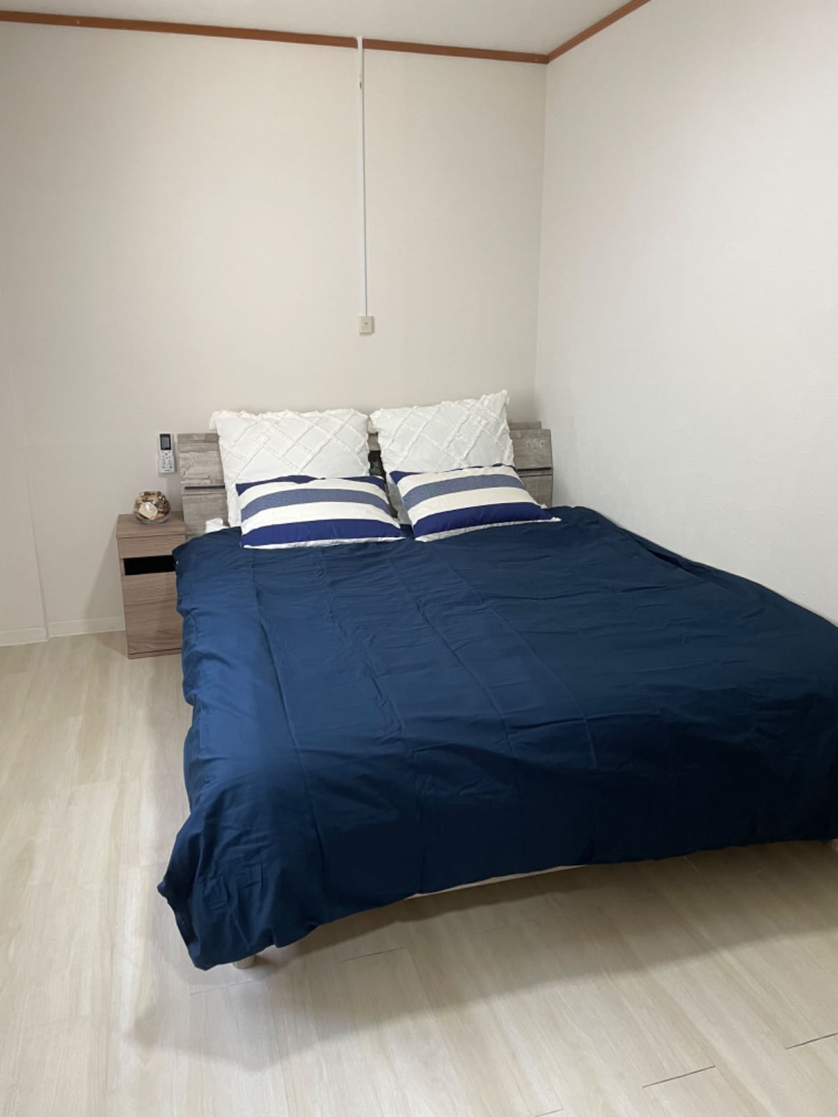 A compact sleeping area features a spacious bed with a navy blue coverlet and two decorative pillows. A bedside table holds a small lamp, while light-colored walls and wooden flooring contribute to a serene ambiance.